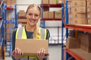 A smiling store associate wearing a high visibility vest holds a box in her hands in the stock room.  