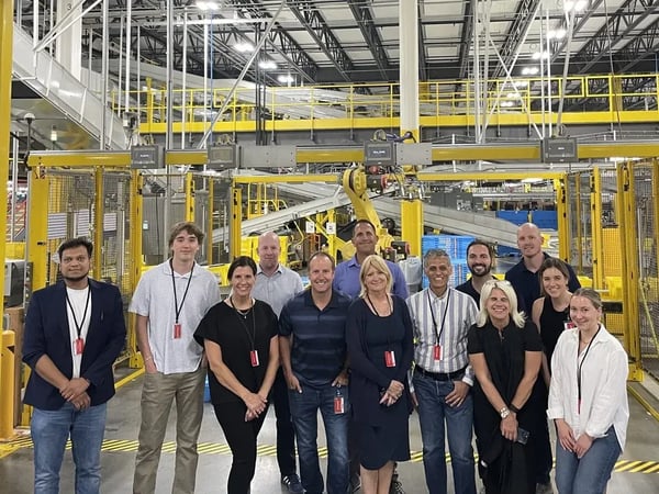 Inside an Amazon Fulfillment Center, Nextuple team members and a large group of retailers pose together.