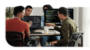A group of four developers huddle around multiple computer screens while planning system integration for a specialty retailer.