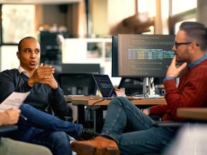 Two developers talk about engineering services beside a desktop displaying code.