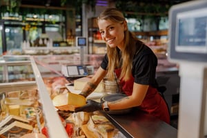 An associate working at the specialty food service counter of a grocery store holds up a wheel of cheese.